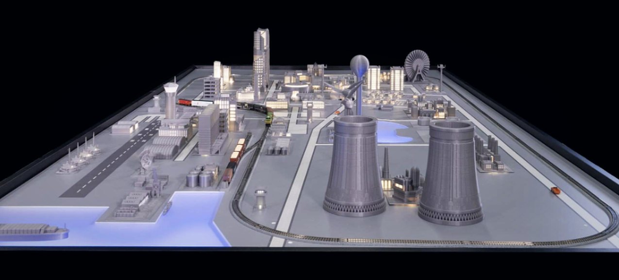 Photo of a city-scale training model with cooling towers, a rail loop, downtown, and a lit harbor